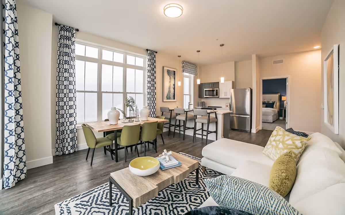 Bright and modern open-concept living space featuring a white sectional sofa with patterned pillows, a wooden coffee table with decorative items, a dining table with green chairs and white vases, large windows with blue and white patterned curtains, and a kitchen area with stainless steel appliances and bar stools. A bedroom is visible through an open doorway.