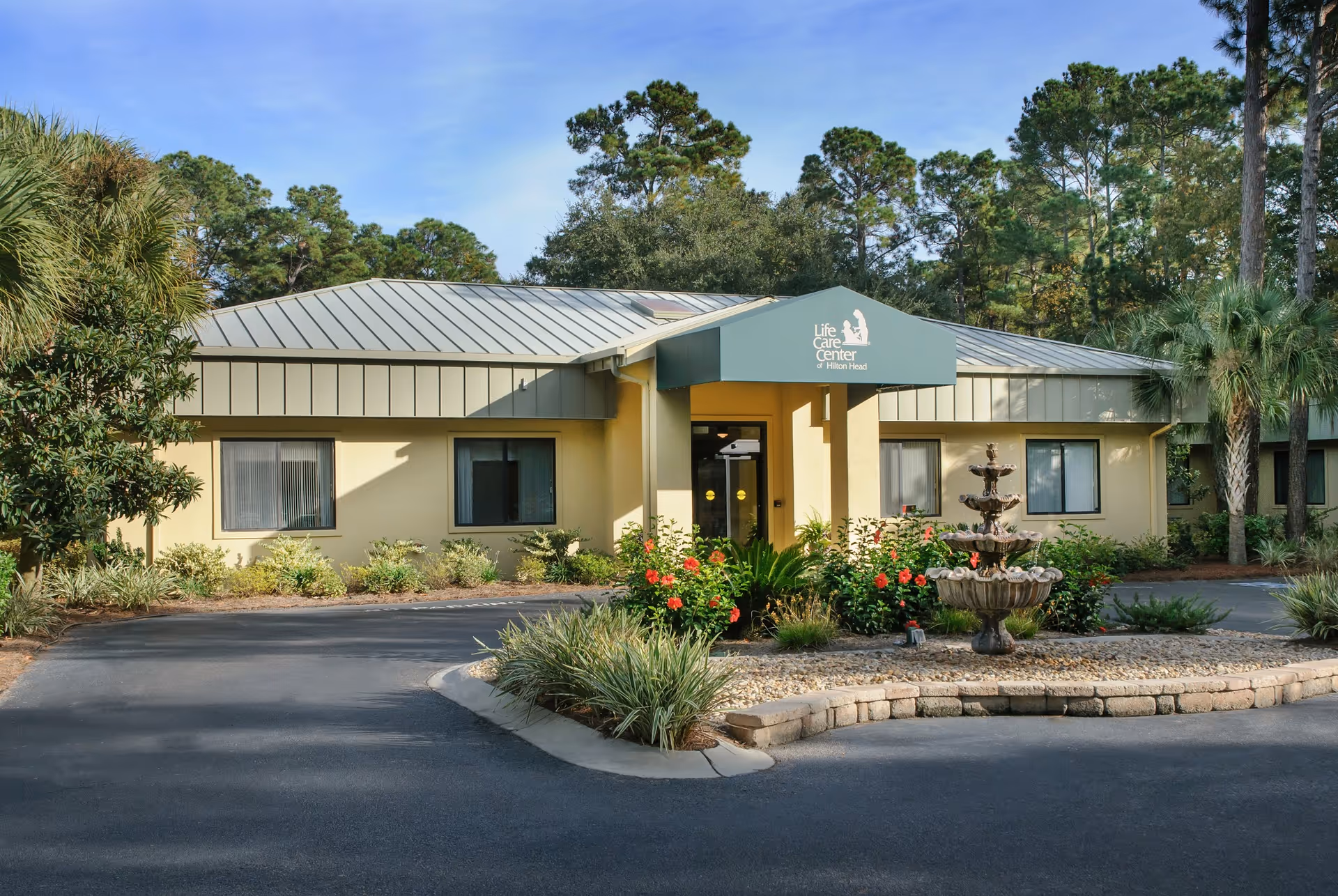 Exterior view of the Life Care Center of Hilton Head building with a green awning over the entrance, surrounded by trees, shrubs, and a decorative fountain in front.