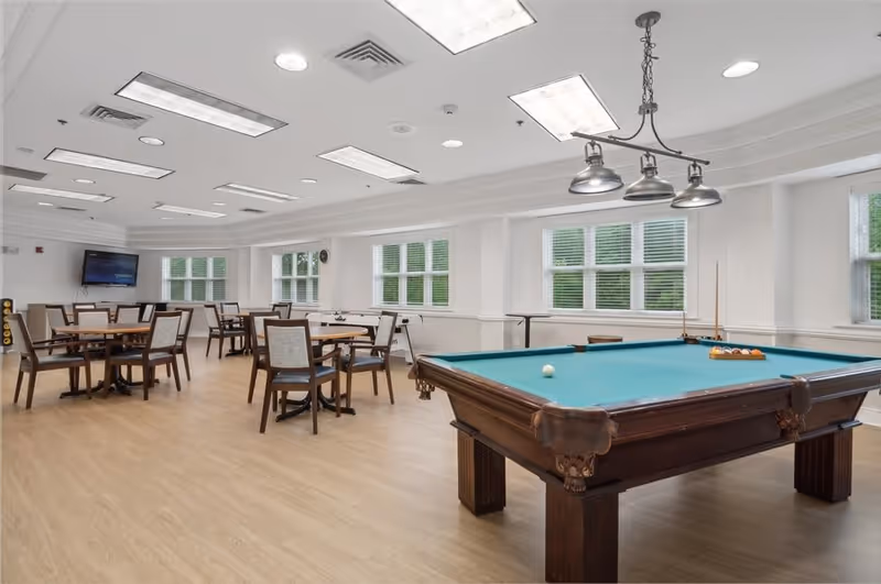 A spacious recreational room with a pool table in the foreground and several round tables with chairs arranged in the background. The room has large windows with white blinds, light wood flooring, and bright overhead lighting. A wall-mounted TV is visible on the far wall.