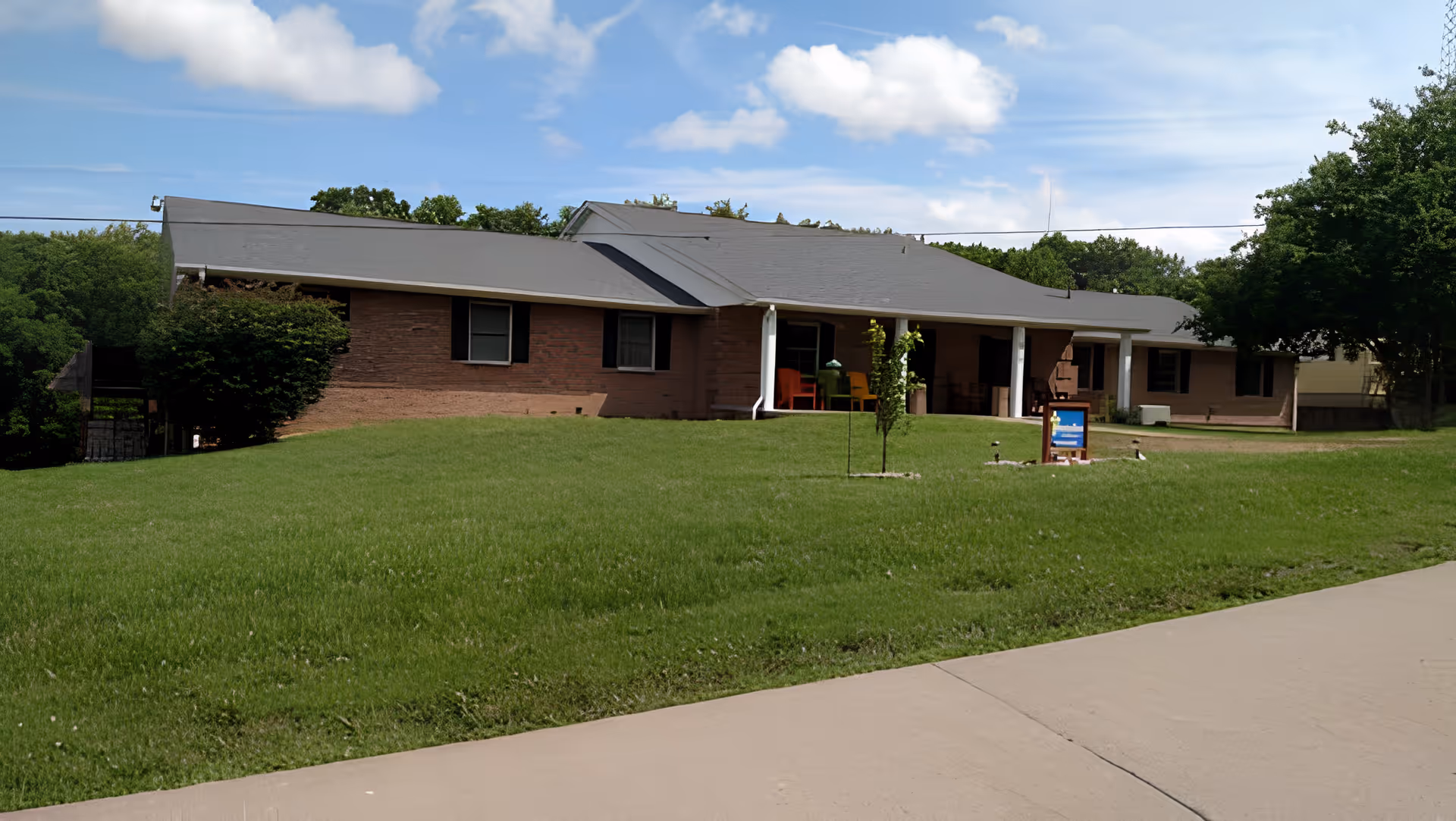 Single-story brick building with a gray roof, surrounded by a well-maintained green lawn and a few small trees, under a partly cloudy blue sky. There is a concrete driveway in the foreground and a small sign near the entrance.
