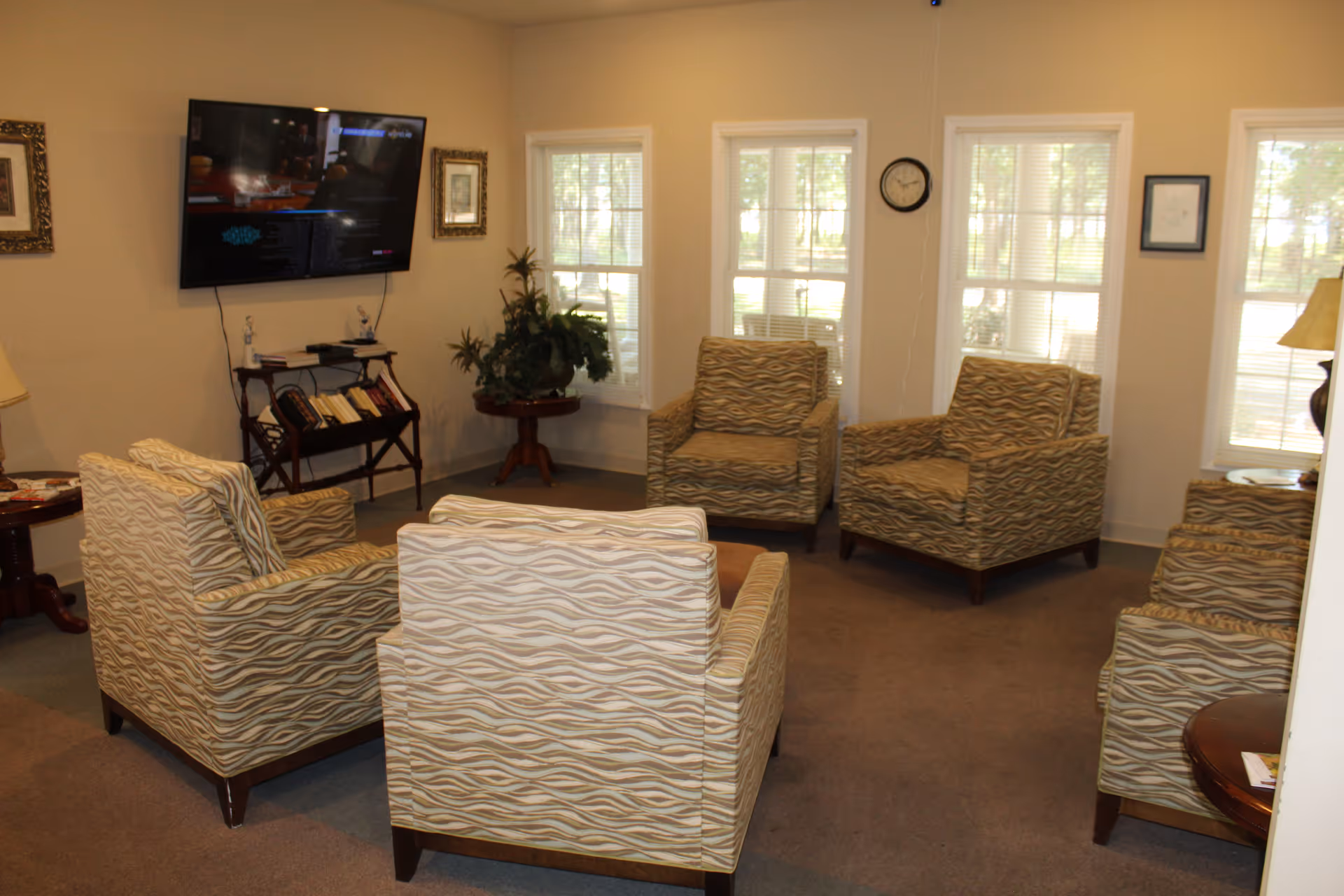 A cozy living room area with six patterned armchairs arranged in a circle on a carpeted floor. The room has several windows letting in natural light, a wall-mounted flat-screen TV, framed pictures on the walls, a clock, and small tables with lamps and plants.