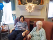 Two elderly women sitting in red armchairs in a cozy living room with a window, lamp, and decorative items on a mantel behind them.