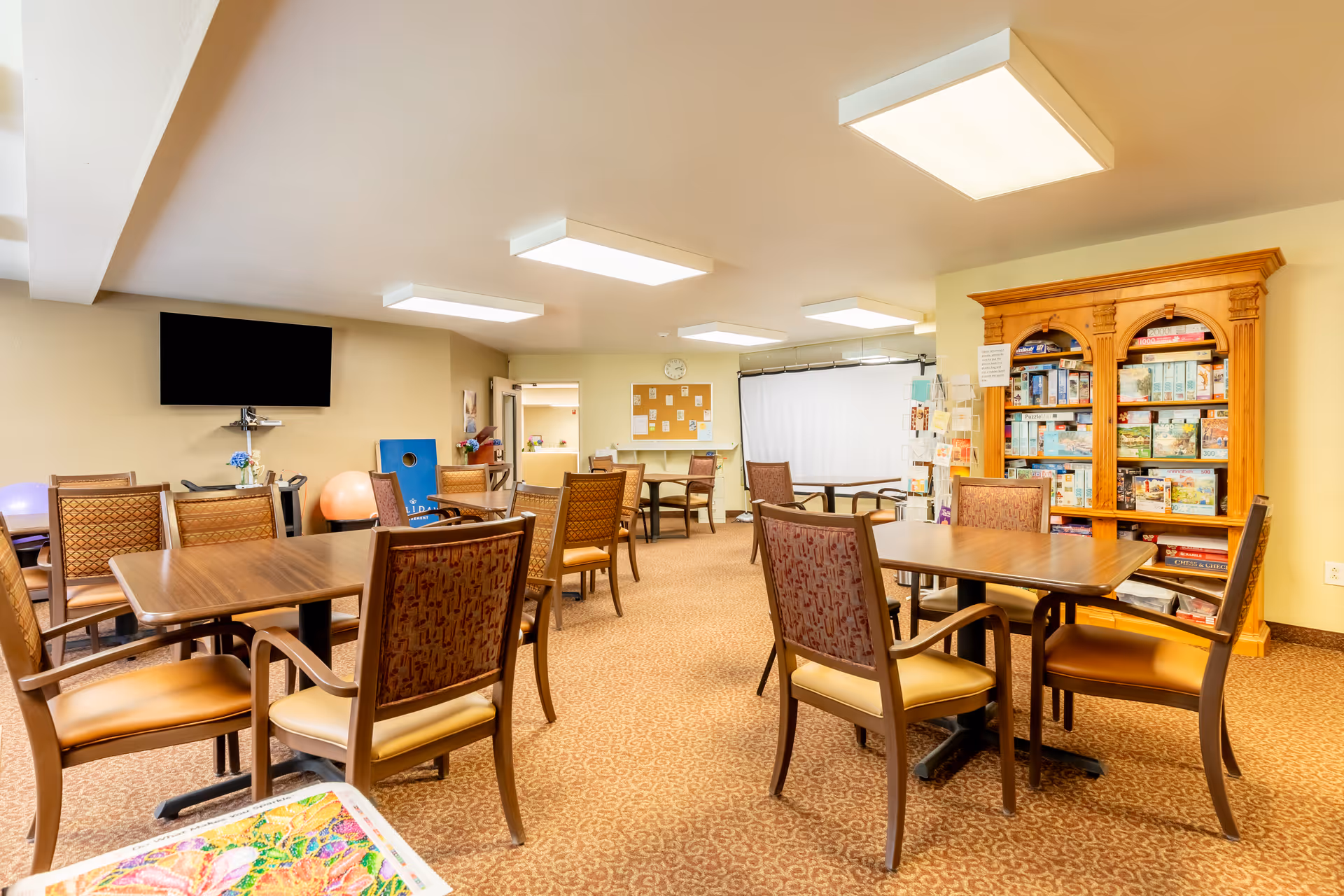 A well-lit common room with multiple wooden tables and cushioned chairs arranged neatly. A large wooden bookshelf filled with board games is visible on the right side. A flat-screen TV is mounted on the left wall above a small table with a flower vase. The room has beige walls and carpeted flooring, with ceiling lights providing bright illumination.