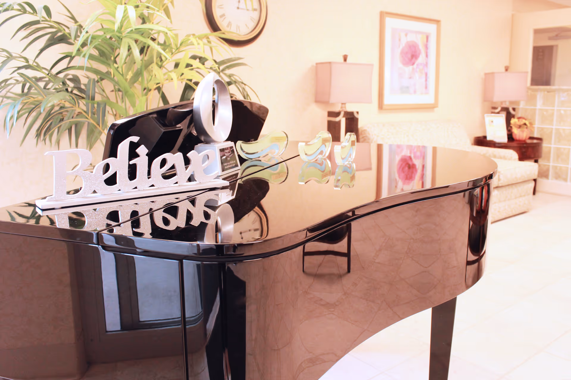 A shiny black grand piano with a decorative 'Believe' sign on top, situated in a warmly lit room with beige walls. In the background, there is a beige sofa, two table lamps with beige shades, a framed floral picture on the wall, a clock, and a large green plant.