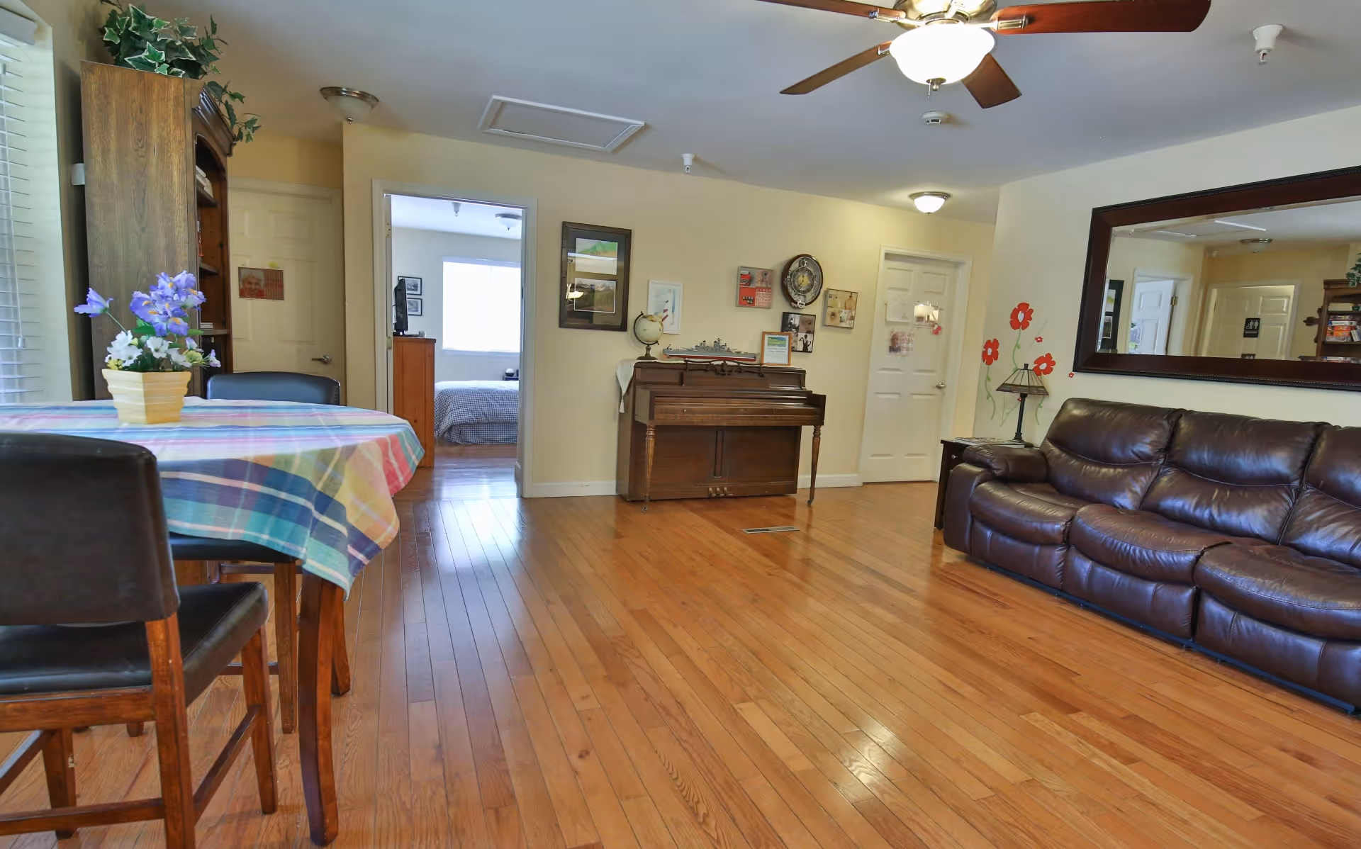 A spacious living room with wooden flooring, a dark brown leather sofa, a large wall mirror, a piano against the far wall, and a small dining table with a colorful checkered tablecloth and a flower pot on it. There are two doorways visible, one leading to a bedroom with a bed and window, and the other to another room. The walls are light-colored with some framed pictures and decorations.