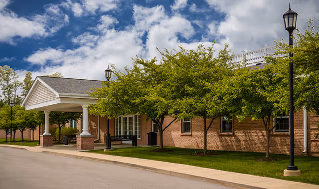 Exterior view of a brick building with a covered entrance supported by white columns, surrounded by green trees and a well-maintained lawn under a partly cloudy sky.