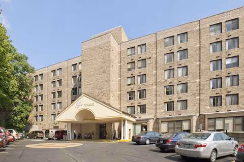 Exterior view of a large brick nursing facility with a porte-cochere entrance and cars.