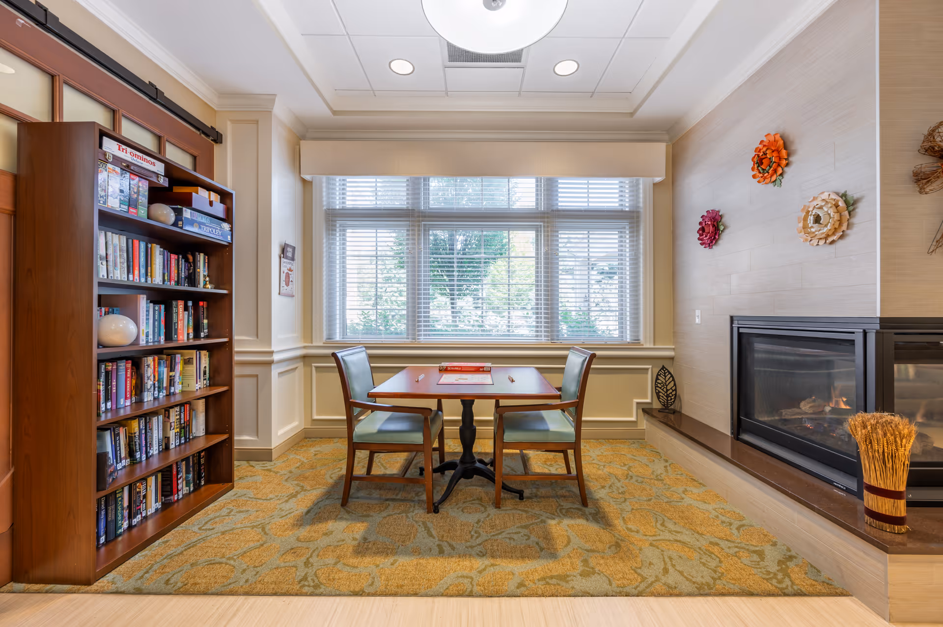 A cozy room with a small wooden table and two chairs placed on a patterned carpet in front of a large window with blinds. To the left, there is a bookshelf filled with books and board games. On the right side, there is a modern fireplace with decorative flowers mounted on the wall above it and a bundle of wheat placed on the hearth.