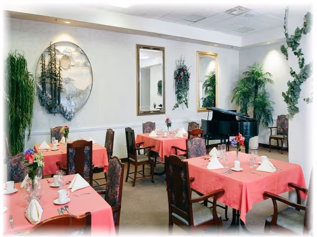 A dining room with several tables covered in pink tablecloths, each set with white napkins, cups, glasses, and small flower arrangements. The room has upholstered chairs, a black grand piano in the corner, large mirrors on the wall, and decorative plants.