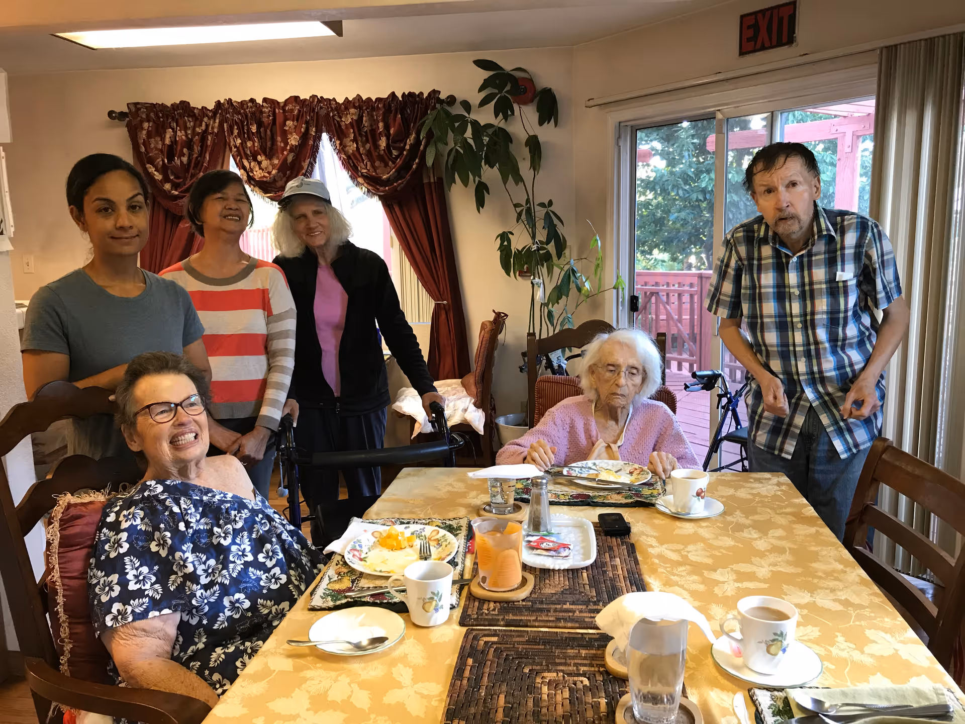 A group of six people gathered around a dining table in a residential care facility. Three elderly individuals are seated at the table with plates of food and cups, while three caregivers or visitors stand behind them. The room has a large window with red curtains and a sliding glass door leading to an outdoor deck. There is a plant in the corner and an exit sign above the door.