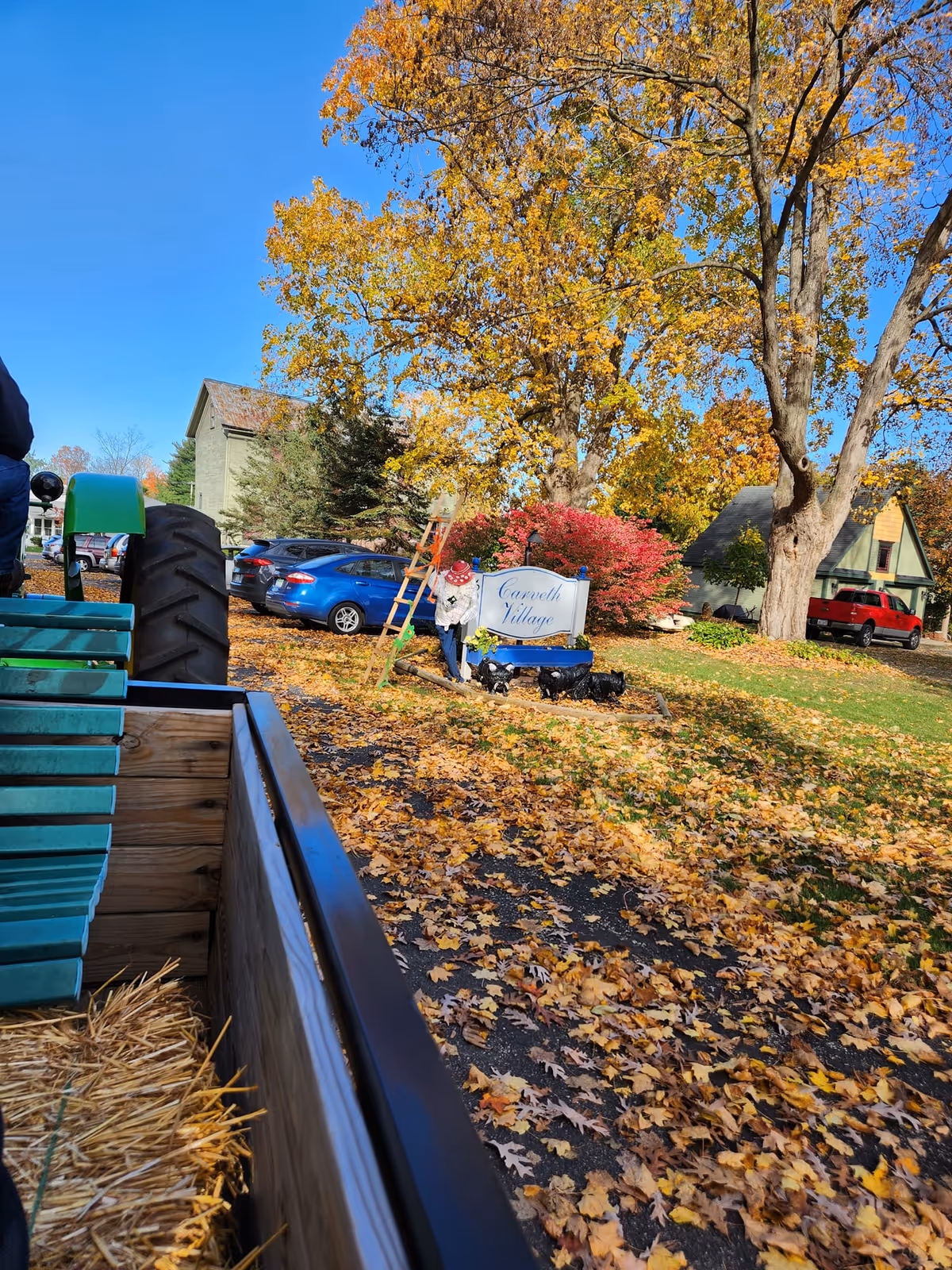Autumn outdoor scene showing the Carveth Village sign under a large tree with parked cars and a hay-filled trailer in the foreground.