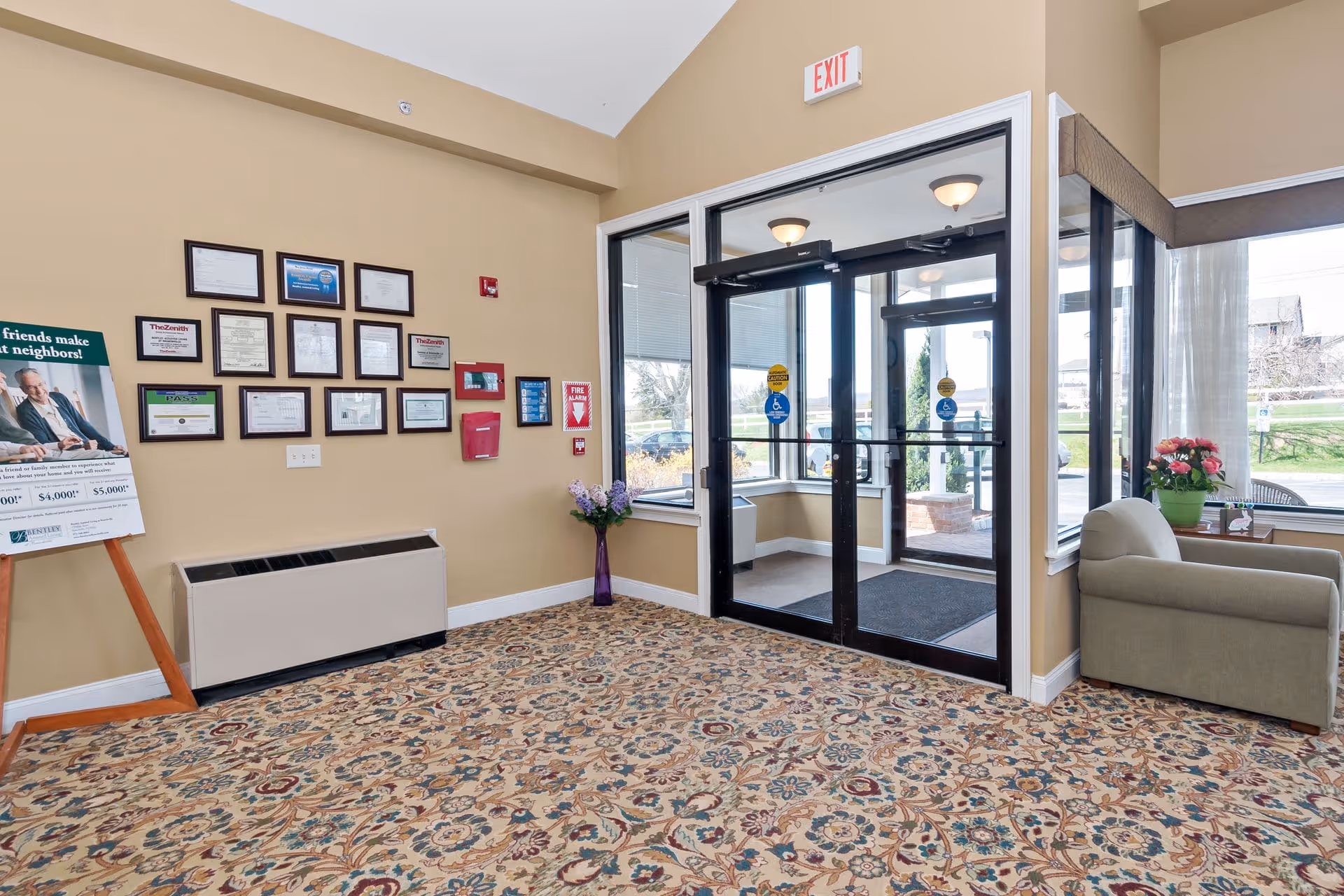 Entrance area of Bentley Assisted Living at Branchville with glass double doors, a patterned carpet, a beige armchair with a side table holding a flower pot, framed certificates and notices on the wall, a fire alarm, and a promotional sign on an easel.
