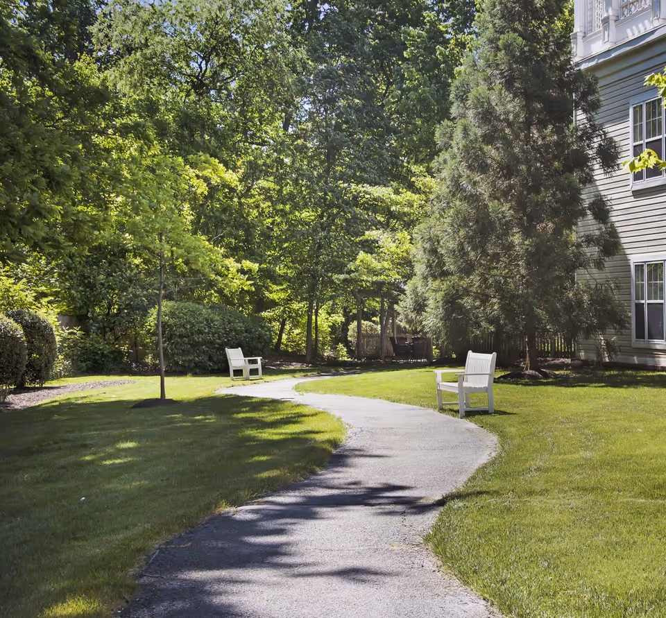 A winding paved path through a grassy courtyard with white benches, trees, and part of a building.