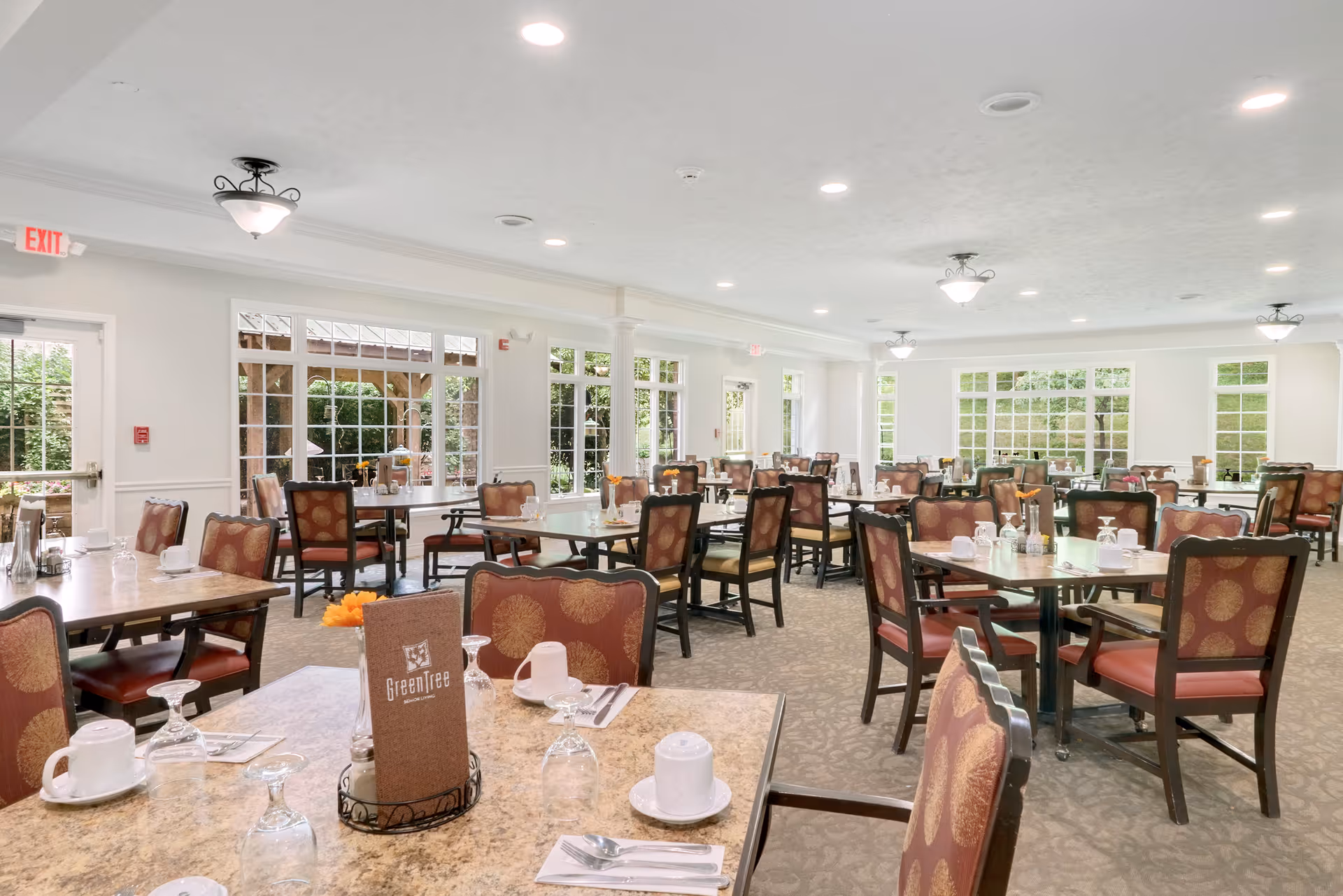 A spacious dining room with multiple tables and chairs arranged neatly. Each table is set with cups, glasses, napkins, and utensils. Large windows allow natural light to brighten the room, and there are ceiling lights evenly spaced across the ceiling. The room has a calm and inviting atmosphere.