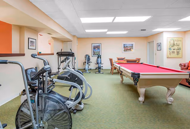 A multipurpose interior room with cardio exercise machines, a red-felt pool table, and seating in a senior living facility.