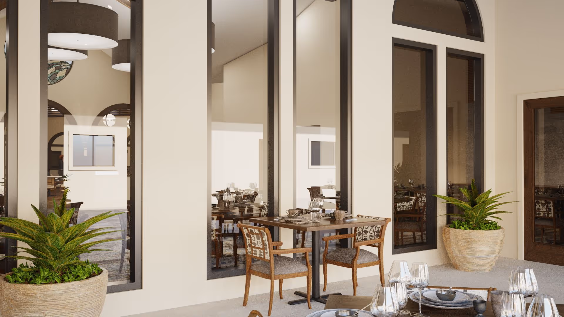 Outdoor patio area with a small dining table set for four, surrounded by wooden chairs with patterned cushions. Large potted plants flank the seating area, and tall windows reflect the interior dining room visible inside the building.