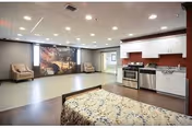 A spacious interior room showing a kitchenette with stove and sink, a bed in the foreground, two armchairs by a large wall mural, and recessed ceiling lights.