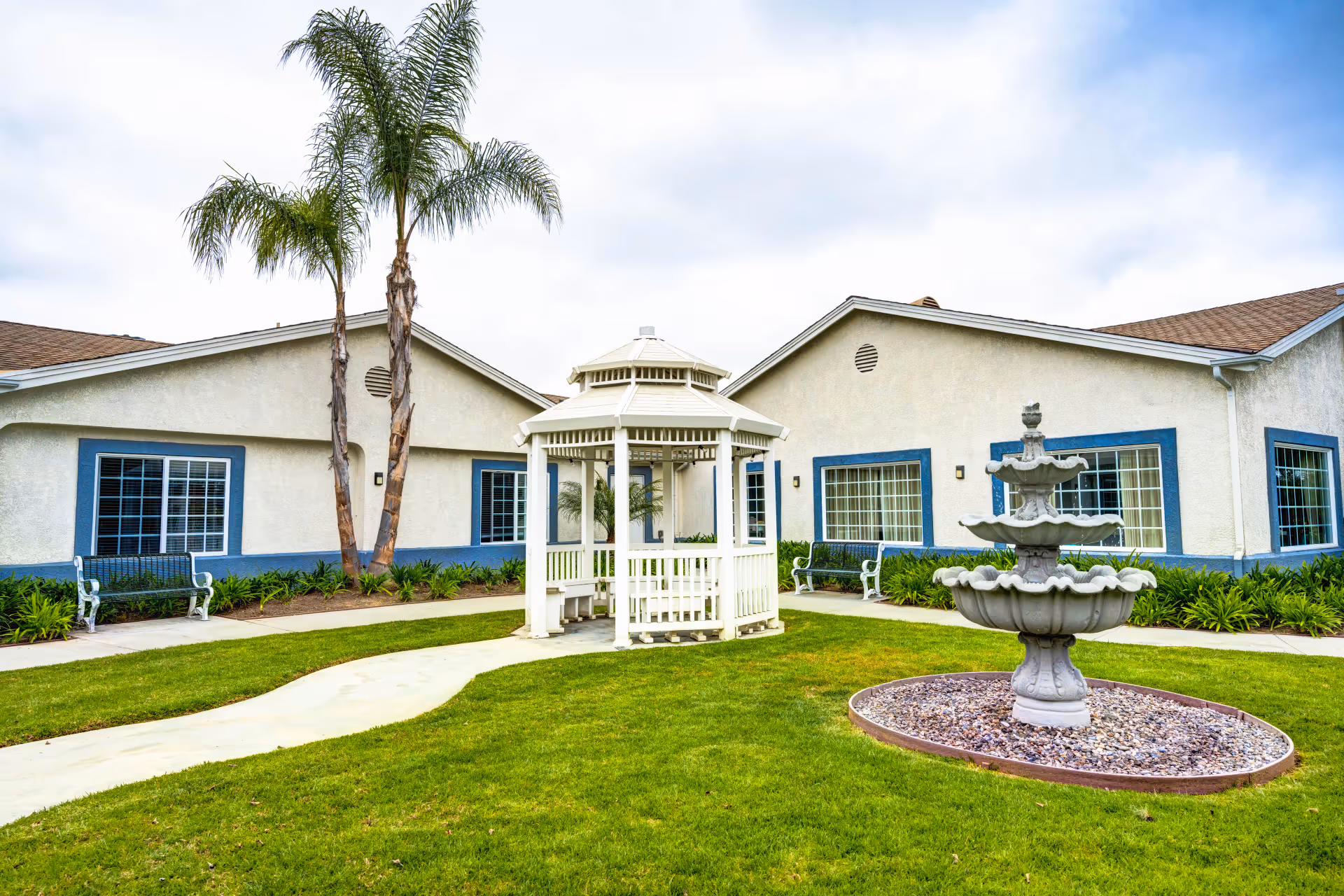 Outdoor garden area at Ivy Terrace at Garden Grove Memory Care featuring a white gazebo, a three-tiered stone fountain surrounded by rocks, green grass, two palm trees, benches, and a beige building with blue window frames in the background under a partly cloudy sky.