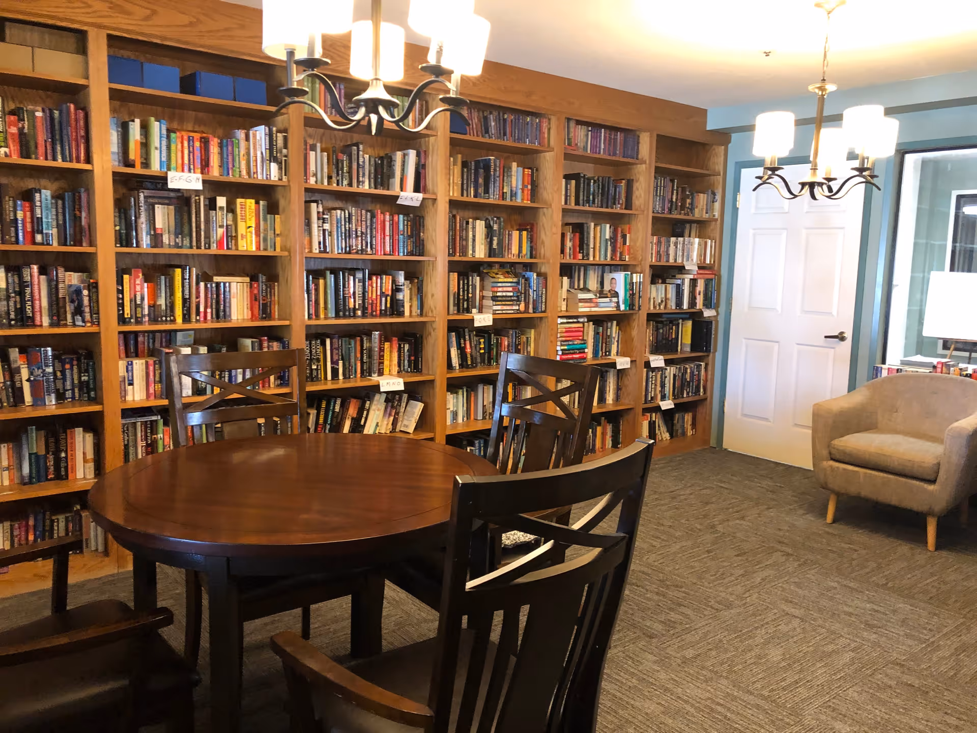 A cozy reading room with wooden bookshelves filled with books lining the walls. In the center, there is a round wooden table surrounded by four wooden chairs. Two chandeliers with multiple lights hang from the ceiling. A beige armchair is placed near a white door and a window with a lamp on a side table is visible in the background.