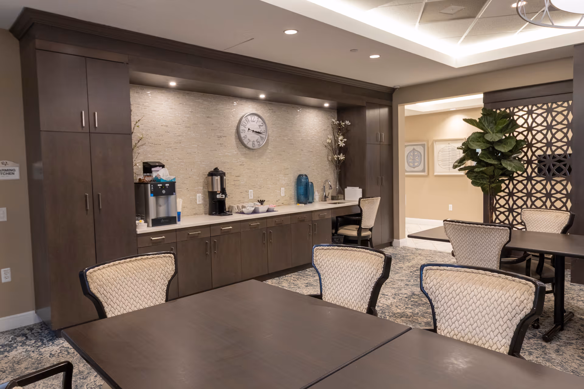 Communal dining area with dark wood tables and upholstered chairs in front of a kitchenette with cabinets, coffee station, and a wall clock.