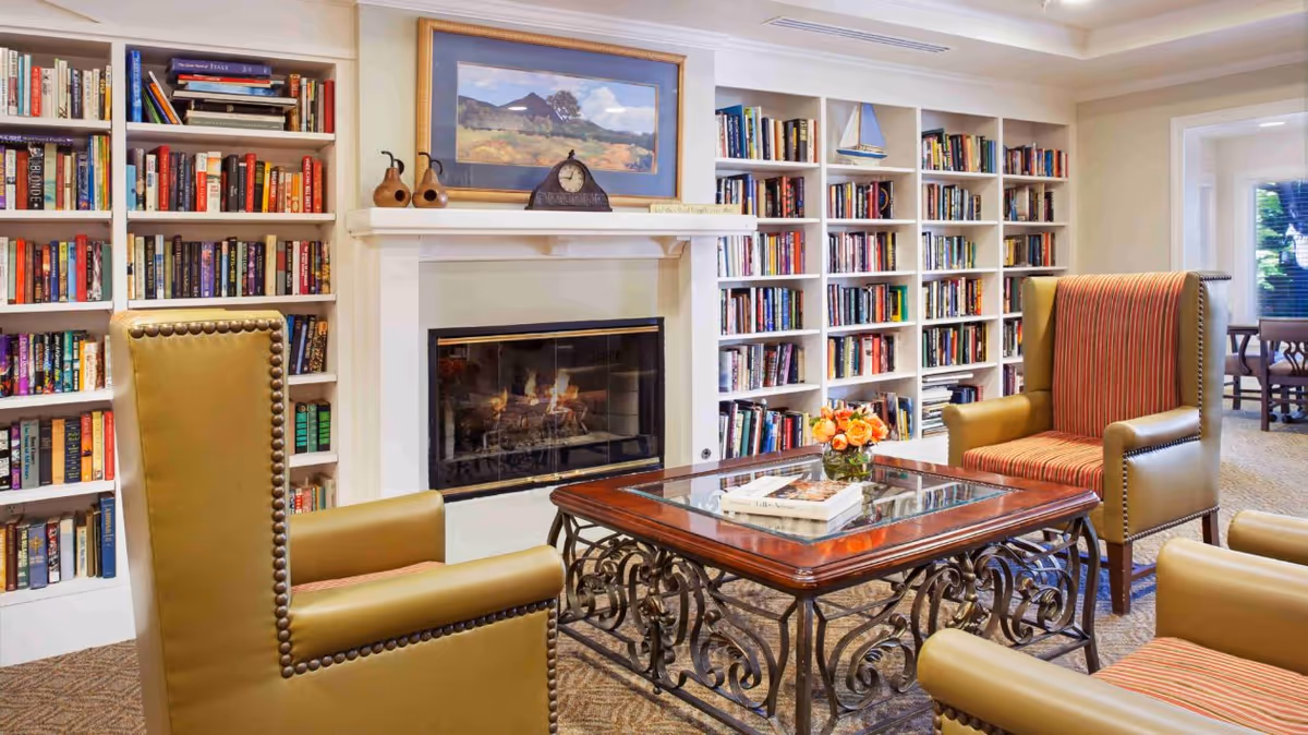 Cozy common room with built-in bookshelves, a fireplace, armchairs and a glass-top coffee table with flowers.