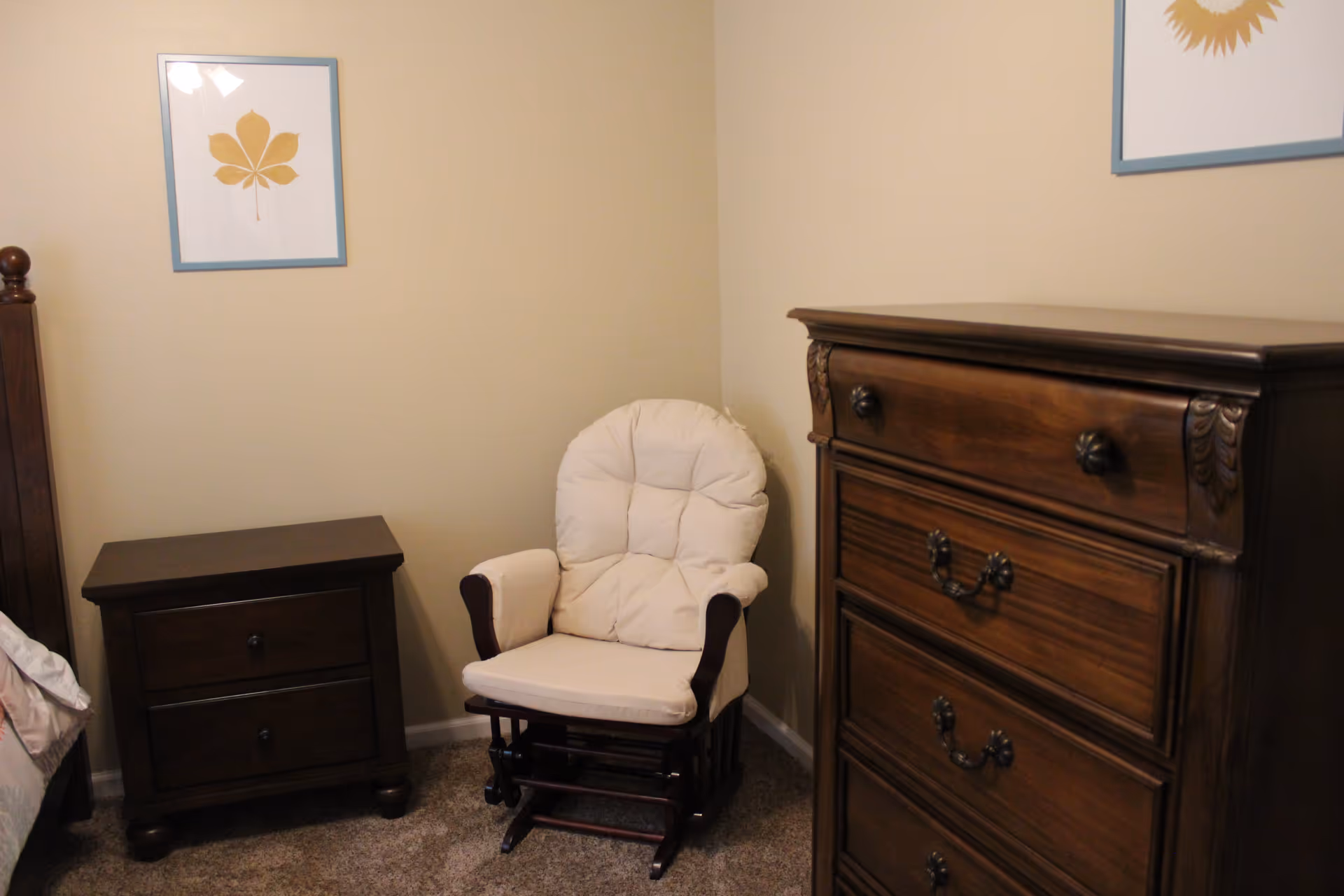 A corner of a bedroom featuring a wooden nightstand with two drawers, a cushioned white rocking chair, and a wooden dresser with four drawers. The walls are beige and decorated with framed artwork of a yellow leaf.
