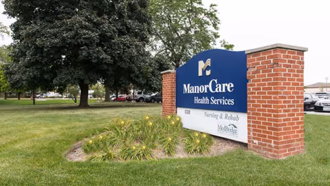 A large blue and white sign for ManorCare Health Services, Nursing & Rehab, and MedBridge, set on a brick base with grass and flowers in front and trees and parked cars in the background.