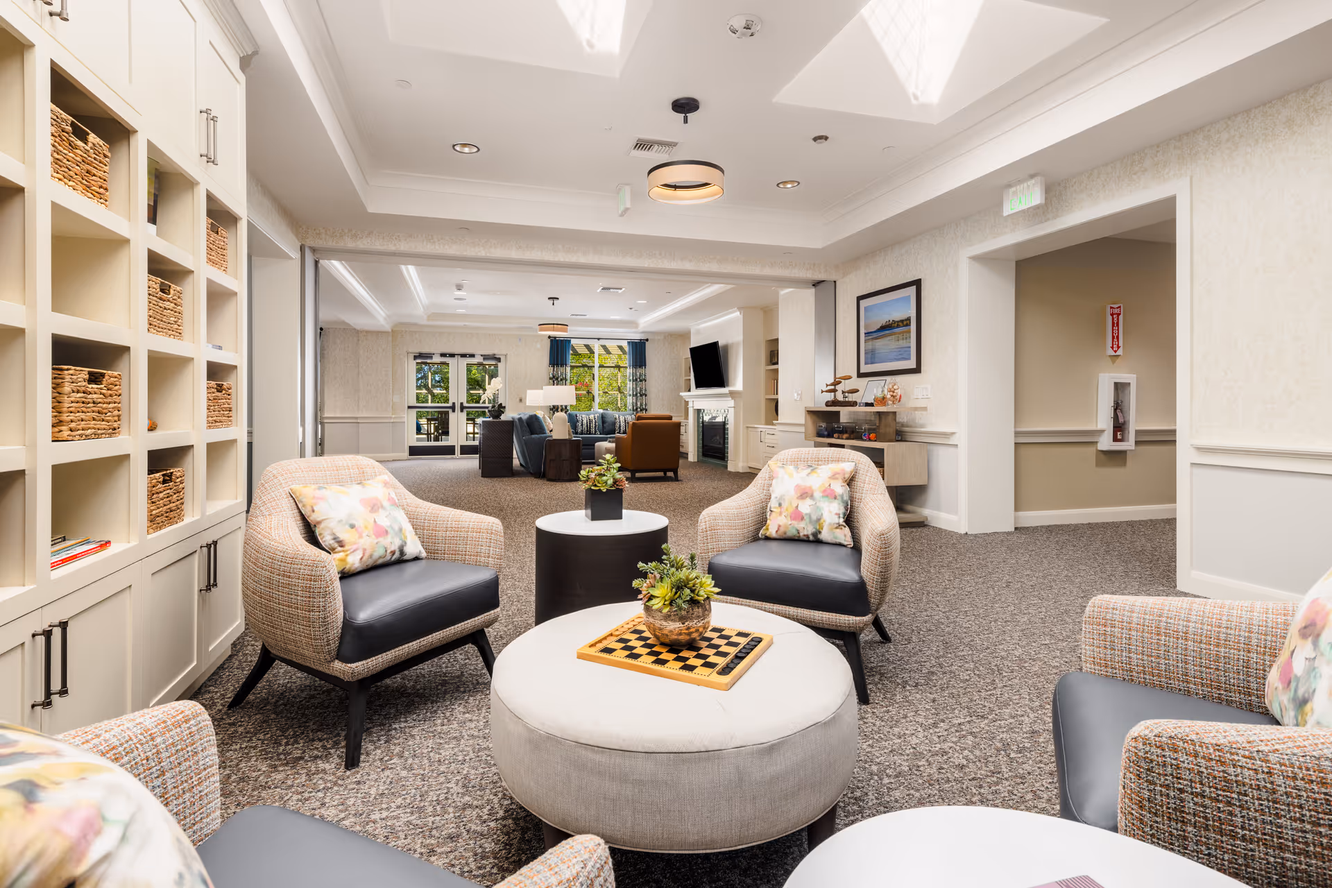 Bright, comfortable senior living lounge with upholstered armchairs around an ottoman topped with a checkerboard, built-in shelving, and a seating area in the background.
