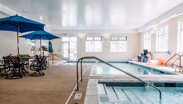 Indoor swimming pool and adjacent hot tub in a bright recreation room with tables, blue umbrellas, and large windows.