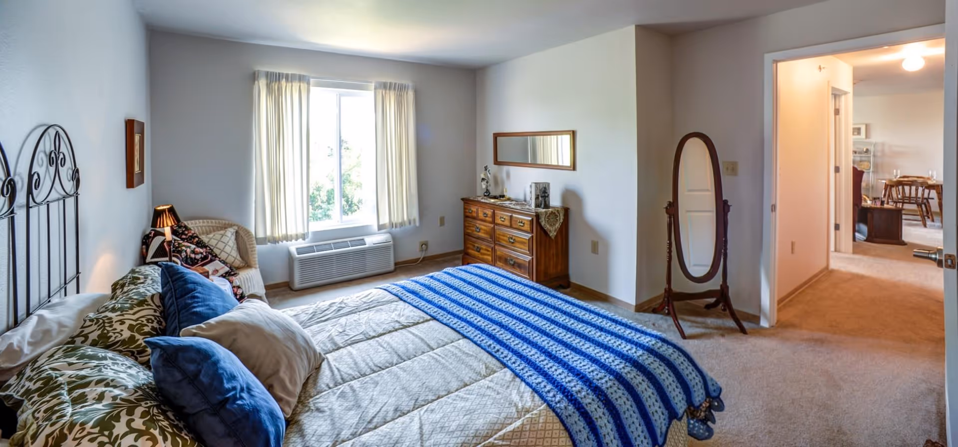 A bright bedroom with a large bed covered in a beige comforter and blue striped blanket. The bed has multiple pillows and a decorative metal headboard. There is a wicker chair with cushions near a window with cream-colored curtains. A wooden dresser with a mirror above it is against the wall, and a full-length standing mirror is positioned near the doorway. The door is open, showing a glimpse of a dining area with a wooden table and chairs.