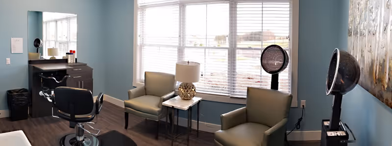 Salon-style room with a styling chair and station, two armchairs by a large window, and hooded hair dryers.