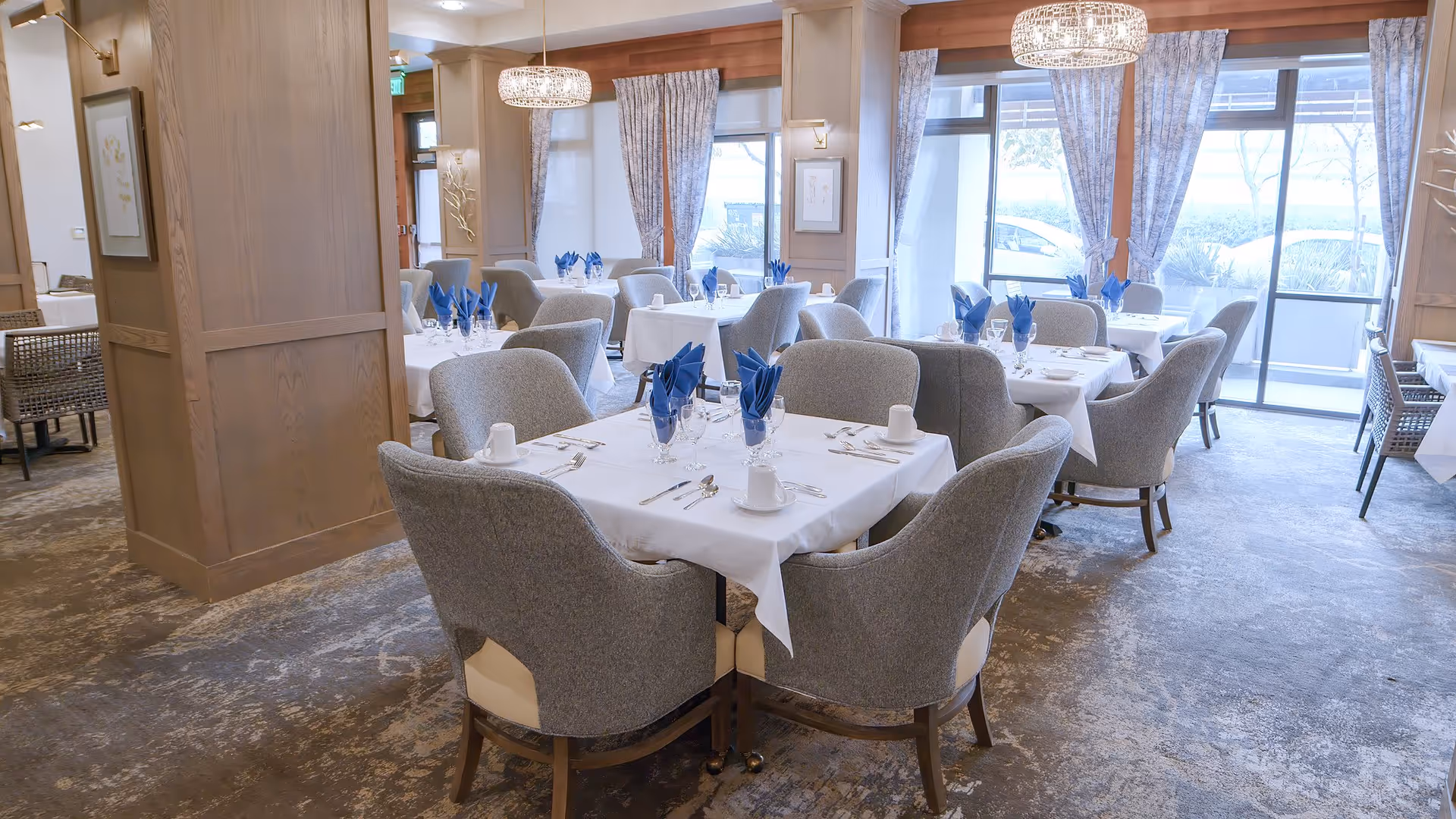 A bright and elegant dining room in a senior living facility with multiple tables covered in white tablecloths, each set with blue folded napkins, glassware, and silverware. Comfortable gray upholstered chairs surround the tables. Large windows with patterned curtains allow natural light to fill the room, and modern chandeliers hang from the ceiling.