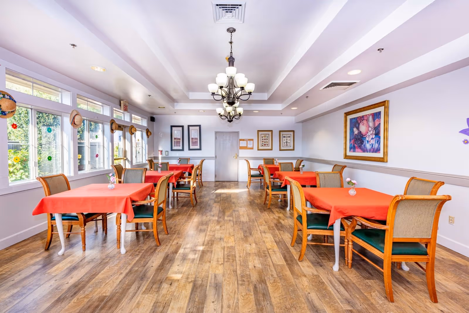 A bright dining room with wooden floors, several tables covered with red tablecloths, each surrounded by wooden chairs with green cushions. The room has large windows on the left side decorated with hats and flower stickers, and a chandelier hanging from the ceiling. Artwork is displayed on the walls.