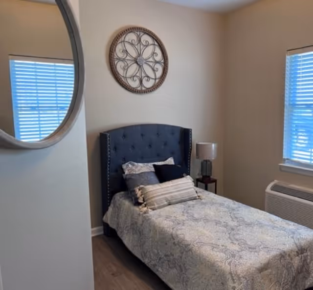 A small bedroom with a single bed featuring a dark tufted headboard, multiple pillows, and a patterned bedspread. There is a round decorative wall piece above the bed, a bedside table with a lamp, a window with blinds, and a wall-mounted air conditioning unit below the window. A round mirror is partially visible on the left wall.