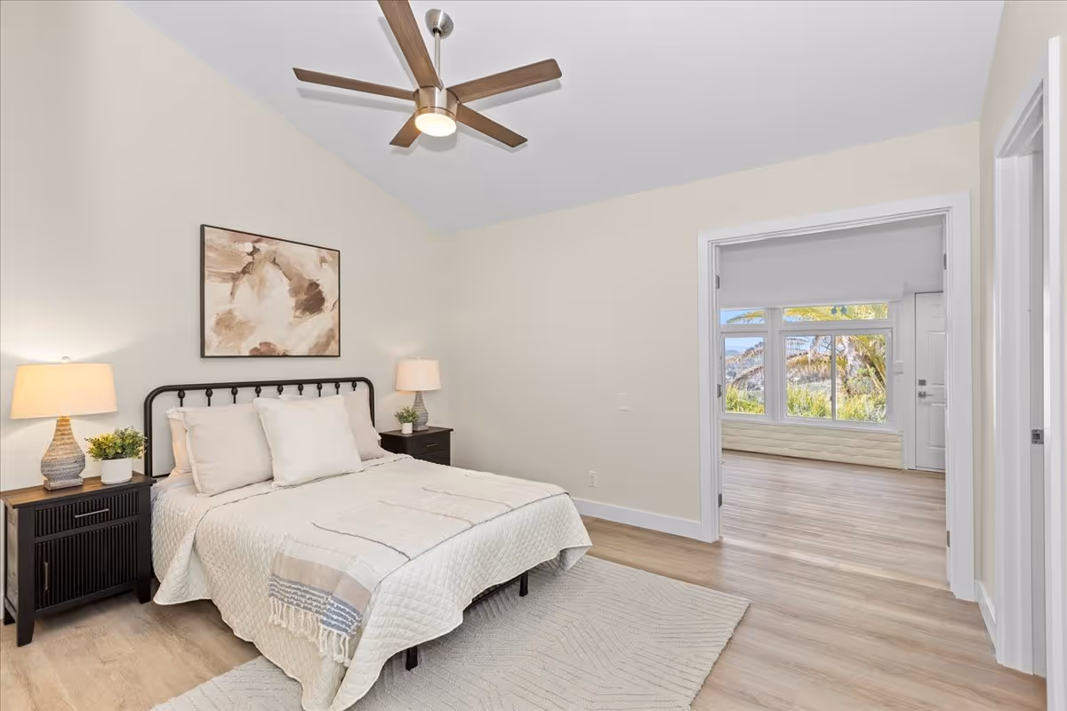 Bedroom featuring a bed with neutral bedding, two nightstands with lamps, a ceiling fan, and an open doorway to a sunlit room with large windows.