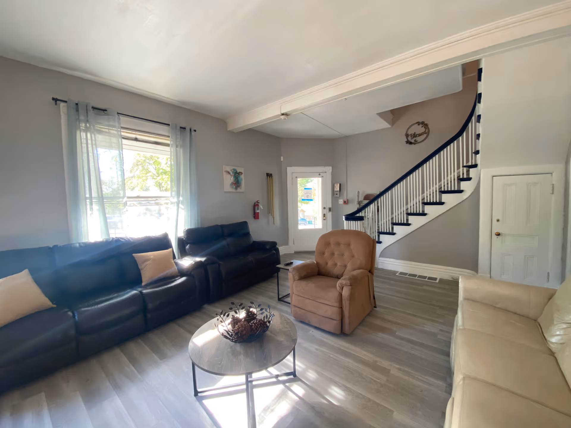 Bright living room with dark leather sofas, a tan recliner, a round coffee table, and a curved staircase by the entry door.