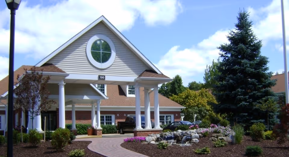 Front exterior view of a senior living facility building with a covered entrance supported by white columns, a circular window above the entrance, landscaped garden with flowers and shrubs, a paved walkway, and a large evergreen tree to the right.
