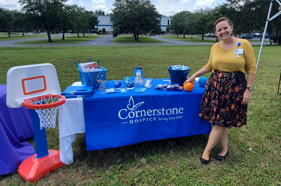 A woman standing outdoors next to a table covered with a blue tablecloth that reads 'Cornerstone Hospice Serving Since 1984.' The table has various items including baskets, pamphlets, and small basketballs. There is a small basketball hoop on the left side of the table. The setting is a grassy area with trees and a building in the background under a partly cloudy sky.