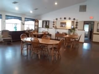 Spacious dining room with several round wooden tables and chairs, hardwood floors, windows, and a serving counter along the back wall.