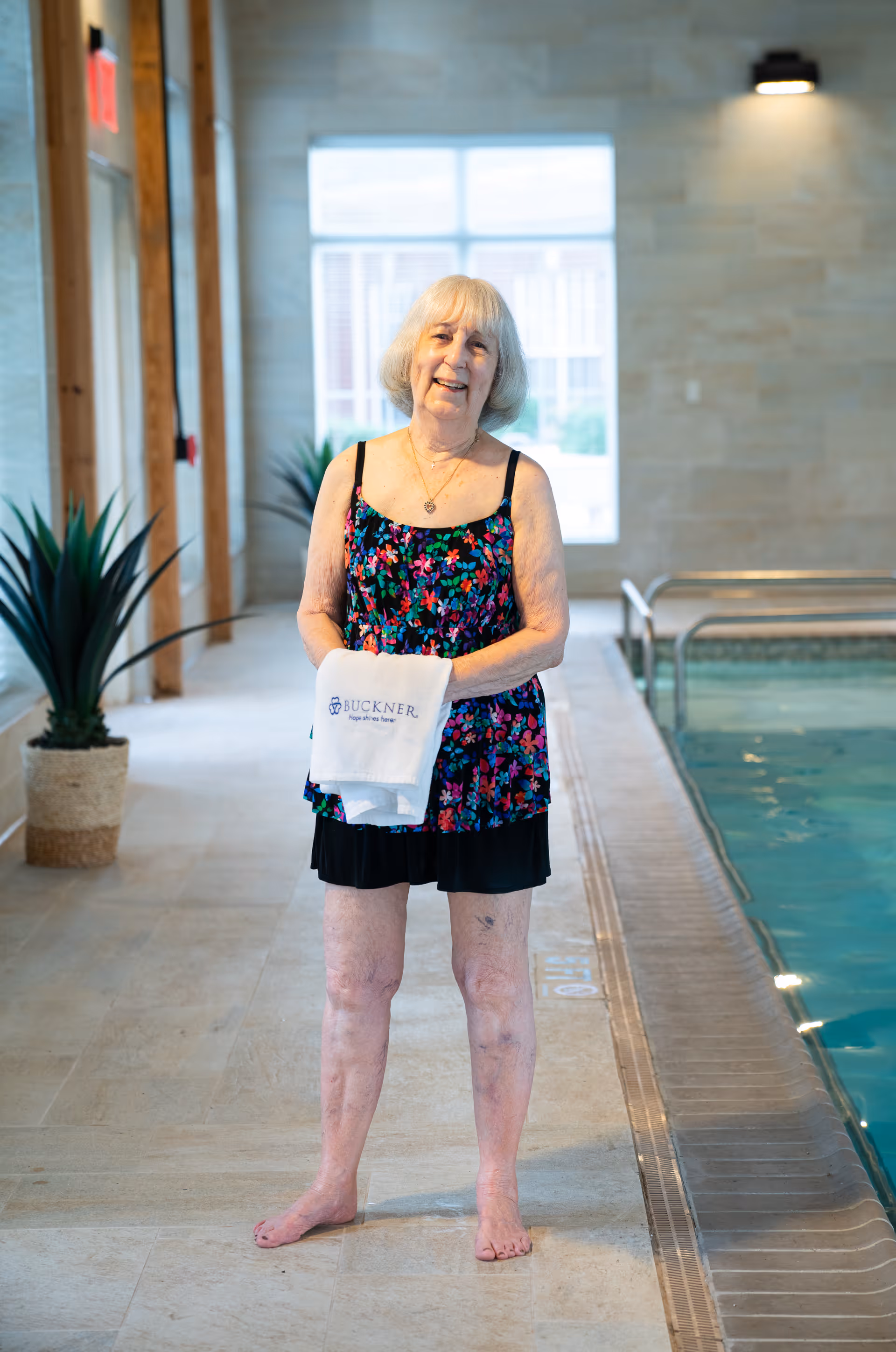 An elderly woman standing barefoot next to an indoor swimming pool, holding a white towel with the Buckner logo. She is wearing a floral swimsuit and smiling. The pool area has tiled floors, potted plants, and large windows letting in natural light.