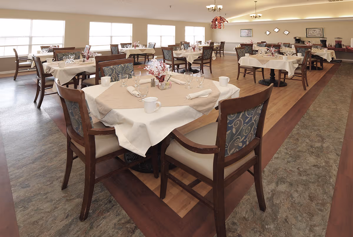 A spacious dining room with multiple tables covered with white and beige tablecloths. Each table is set with cups, glasses, napkins, and a small floral centerpiece. The room has large windows letting in natural light and wooden chairs with patterned upholstery.