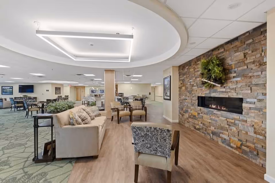 Open senior living common area with sofas, chairs and tables, a modern ceiling light feature and a stone wall fireplace.