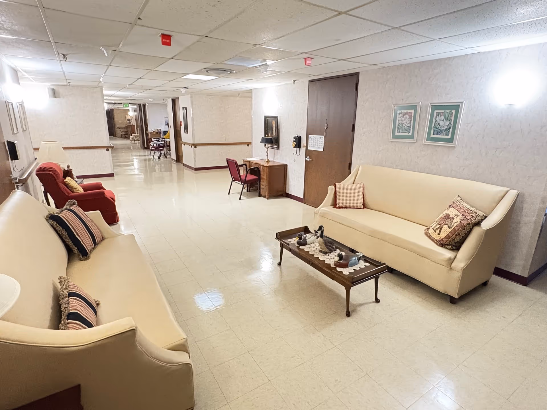 A hallway in a senior living facility with two beige sofas facing each other, each adorned with decorative pillows. A small wooden coffee table with duck figurines is placed between the sofas. There is a red armchair and a small desk with a chair against the wall. The walls are light-colored with framed artwork, and the floor is tiled. The hallway extends into the distance with more furniture visible further down.