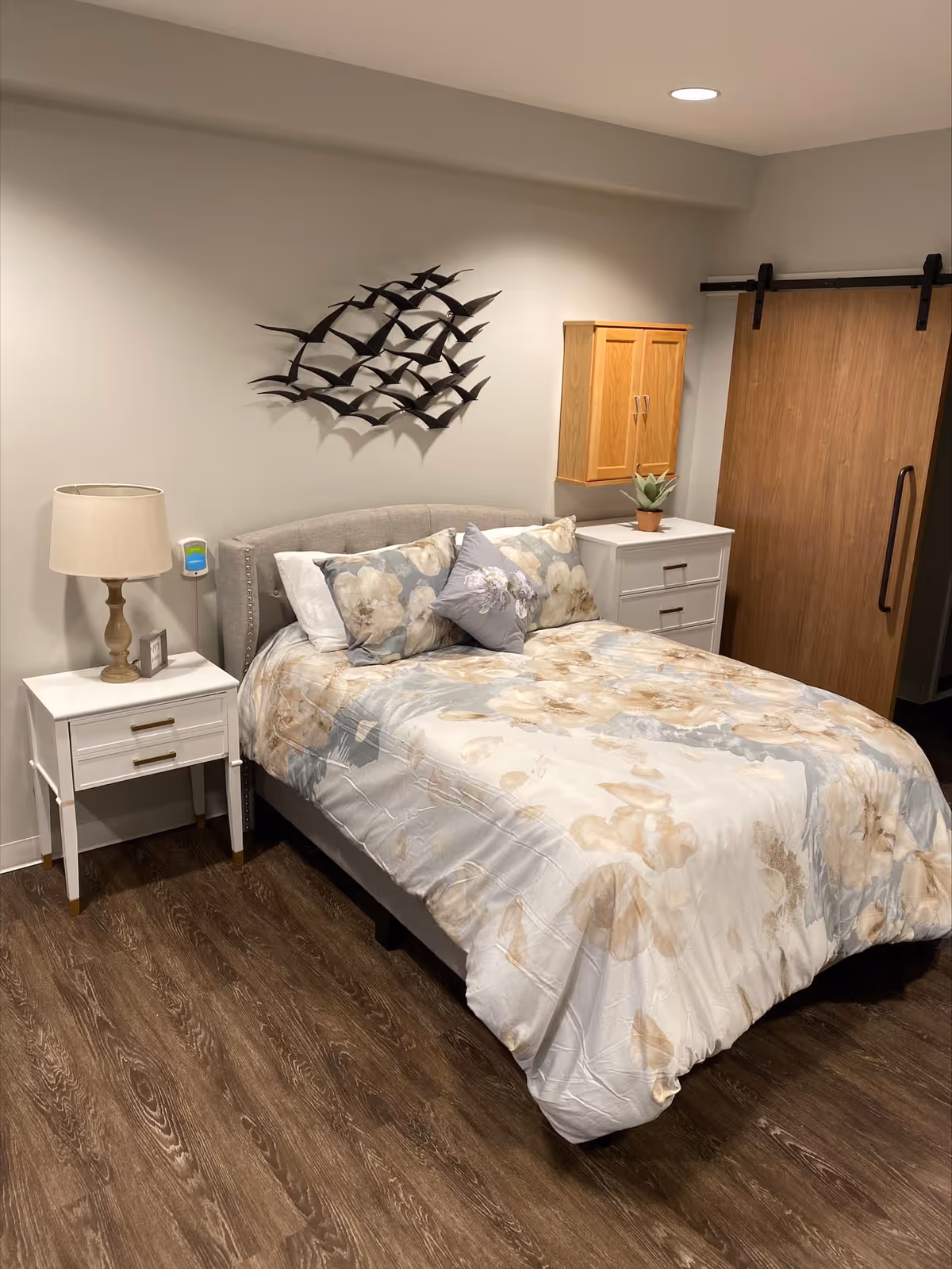 A cozy bedroom with a bed featuring floral-patterned bedding and multiple pillows. There is a white nightstand with a lamp and a small picture frame on the left side of the bed. On the right side, there is a white dresser with a small potted plant and a wooden wall cabinet above it. A decorative metal wall art of birds in flight is mounted above the bed. The room has wooden flooring and a sliding wooden barn door on the right side.