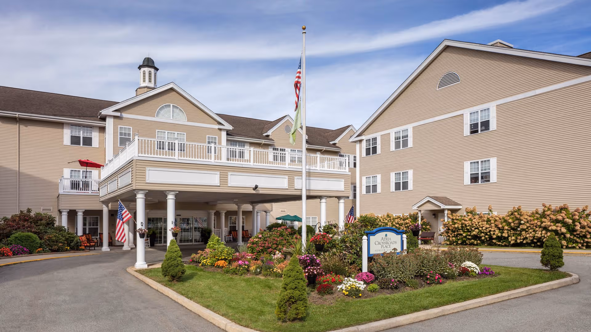 Exterior view of Atria Crossroads Place, a senior living facility with beige siding, white trim, and multiple windows. The entrance features a covered drop-off area supported by white columns. There is a landscaped garden with colorful flowers and shrubs in front of the building, along with a flagpole displaying the American flag. A sign with the facility's name is visible in the garden.
