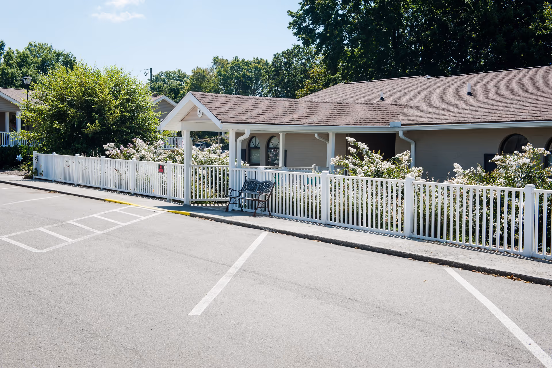 Exterior view of a single-story building with a brown shingled roof and light-colored walls, surrounded by a white fence with flowering bushes behind it. There is a small covered entrance with a bench outside, and a parking lot with marked spaces in the foreground. Trees and greenery are visible in the background under a clear blue sky.