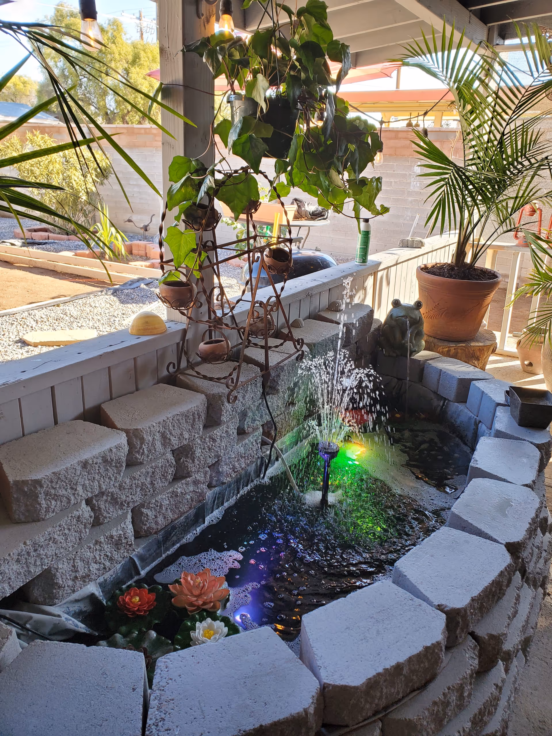 A small indoor water fountain surrounded by light gray stone blocks with colorful LED lights illuminating the water. There are artificial flowers floating in the water and a small frog statue near the back. The fountain is located under a covered patio area with potted plants and hanging greenery, with an outdoor garden visible in the background.