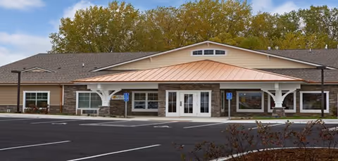 Front exterior of a single-story senior living facility with a covered entrance supported by white posts, stone facade accents, and an empty parking lot.