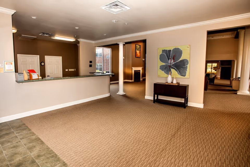 Interior view of a senior living facility reception area with a long counter, two white doors behind the counter, a small table with decorative vases and a large flower painting on the wall, beige carpet flooring, and a hallway leading to a lounge area with seating and a fireplace.