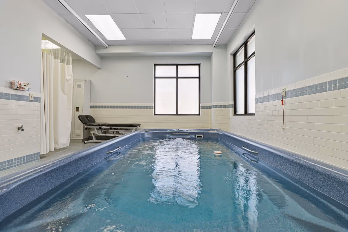 Indoor therapy pool in a tiled rehabilitation room with windows and a treatment table.
